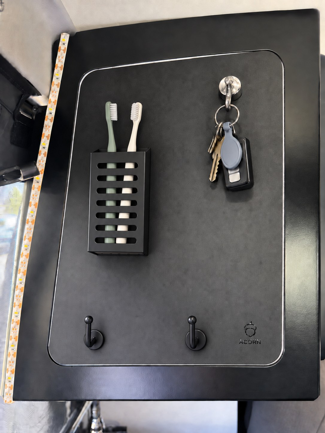 Matte black Acorn Stash Entrance Magnetic Board mounted in the trim configuration of a Winnebago Revel pantry model, holding a slotted black toothbrush holder with two toothbrushes, a chrome magnetic hook with a key ring and fob, and two black J-hooks along the bottom edge.