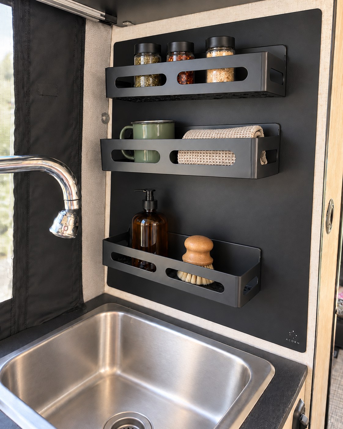 Matte black Acorn Stash Kitchen Magnetic Board mounted beside the stainless steel sink in a Winnebago Revel pantry model, holding three black magnetic shelves with glass spice jars, a green enamel mug, a dish towel, an amber soap dispenser, and a wood dish brush.