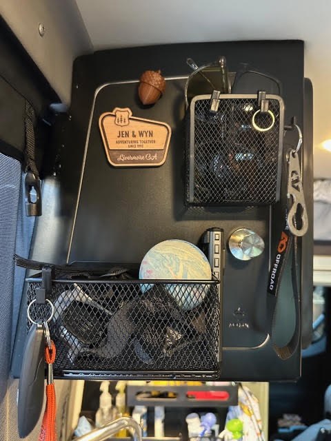 Customer photo from Jenny Penny showing a black Acorn Stash Entrance Magnetic Board in a Winnebago Revel holding an upper wire mesh basket with sunglasses, a lower wire mesh basket of small gear, a carabiner, keys on a red lanyard, and a wooden "Jen & Wyn" name magnet.