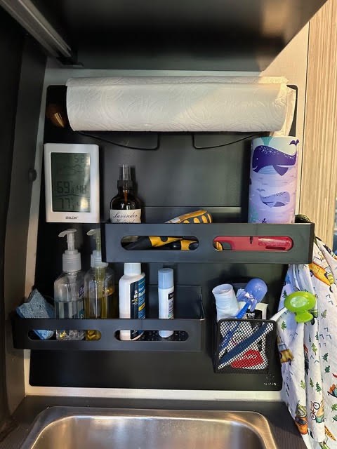 Customer photo from Jenny Penny showing a matte black Acorn Stash Kitchen Magnetic Board behind a Winnebago Revel sink holding a paper towel roll, digital thermometer, lavender spray, utility tools, soap bottles, a sponge, and toothbrushes.